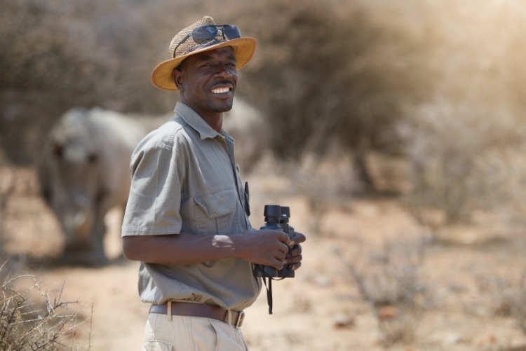 Portrait, safari and wildlife with a man ranger outdoor in a game park for nature conservation. Animals, binoculars and blurred background with an african male person on patrol in the wilderness